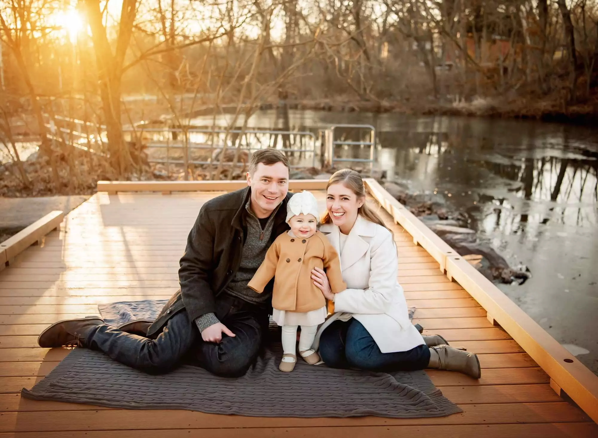 mom and dad smiling with their baby girl by creek sitting on peer at fischers park Lansdale Family and Newborn Photographer