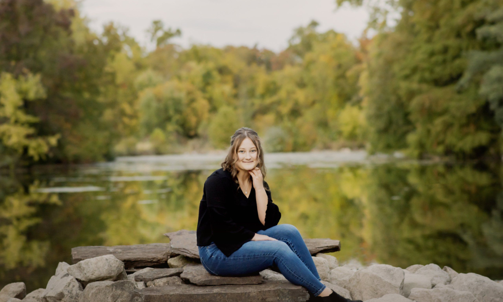 A high school senior sitting on rocks by the water at prophecy creek park at the manor house in ambler