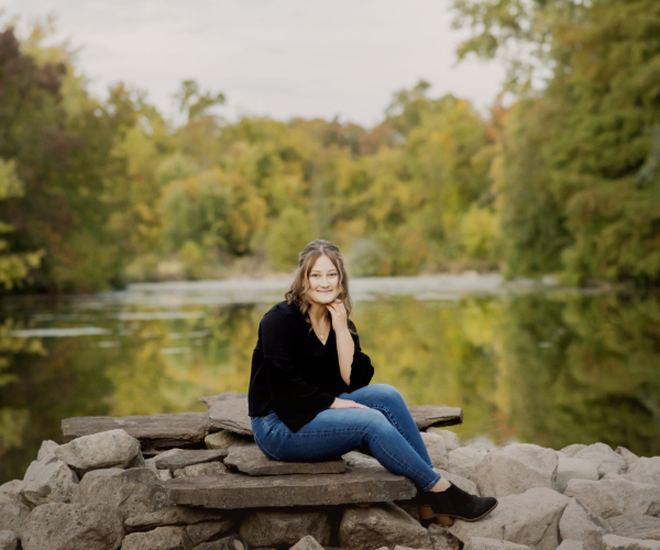 A high school senior sitting on rocks by the water at prophecy creek park at the manor house in ambler