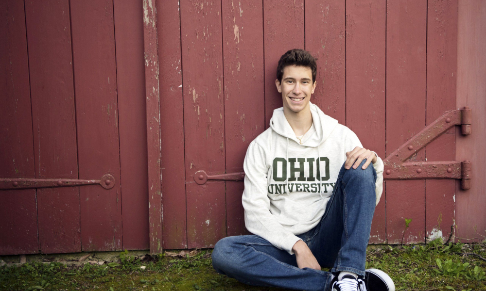 High School Senior sitting in front of red barn wearing Ohio University shirt at Highland Mansions in Ft. Washington Lansdale family and newborn photographer