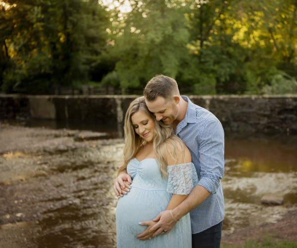 A man in a sky blue check shirt and his pregnant wife in sky blue dress both are looking down a baby in belly fischers park lansdale