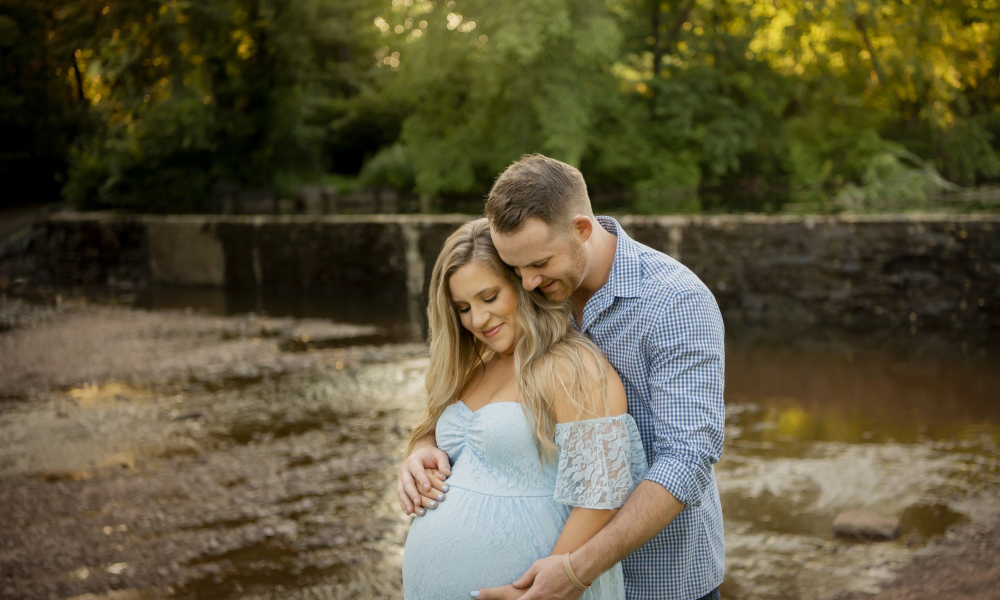 A man in a sky blue check shirt and his pregnant wife in sky blue dress both are looking down a baby in belly fischers park lansdale