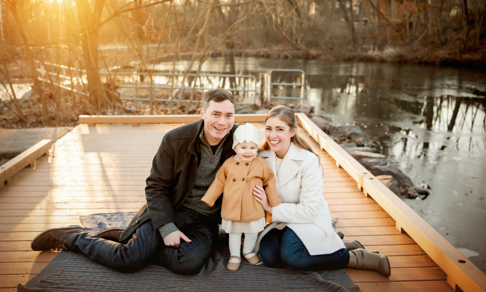 mom and dad smiling with their baby girl on the peer at sunset by the creek at fischers park in Lansdale