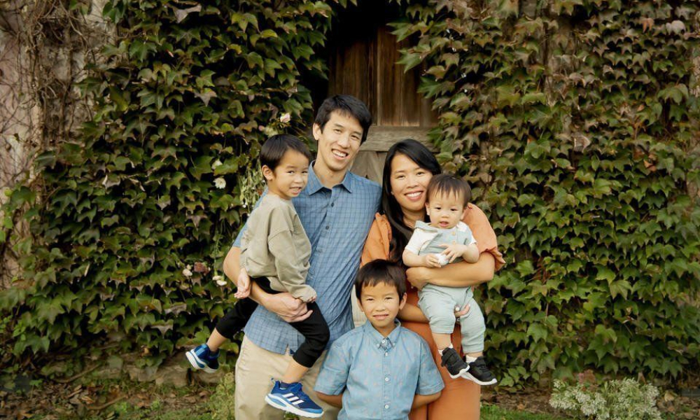 Husband and wife to took 2 boys in the lap and the third one is standing on the ground.