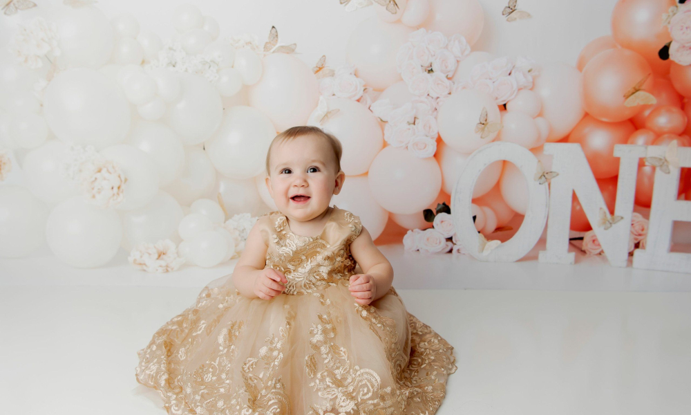 baby girl wearing gold dress smiling on first birthday one peach butterfly balloons Lansdale Family and Newborn Photographer