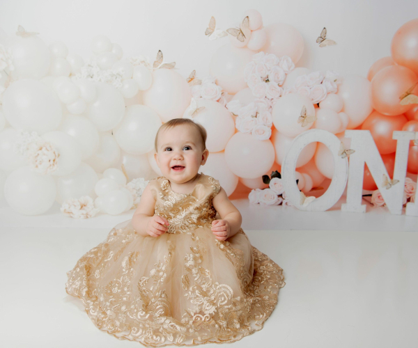 baby girl wearing gold dress smiling on first birthday one peach butterfly balloons Lansdale Family and Newborn Photographer