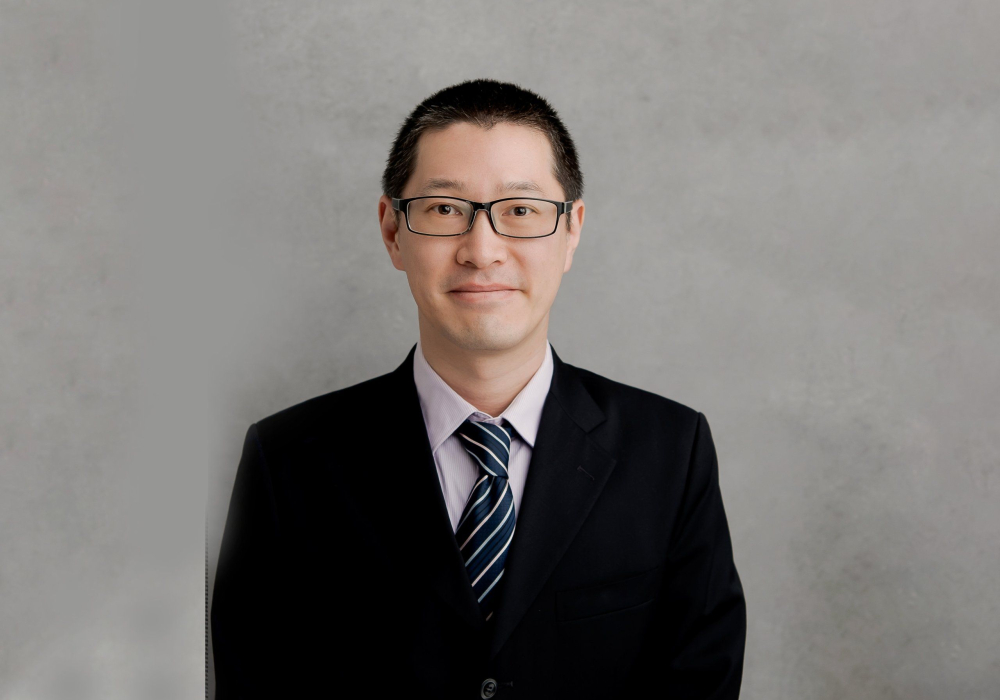 Professional headshot of a men wearing glasses in lavender dress shirt and black jacket