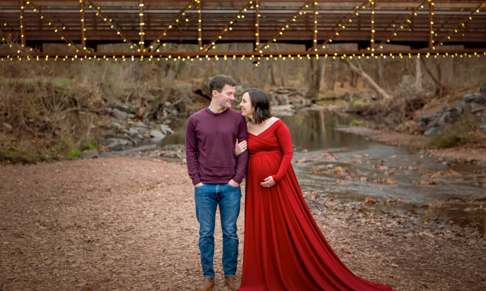 Maternity portraits of husband and wife at Fischer's Park. Mom wearing red dress in front of lighted bridge